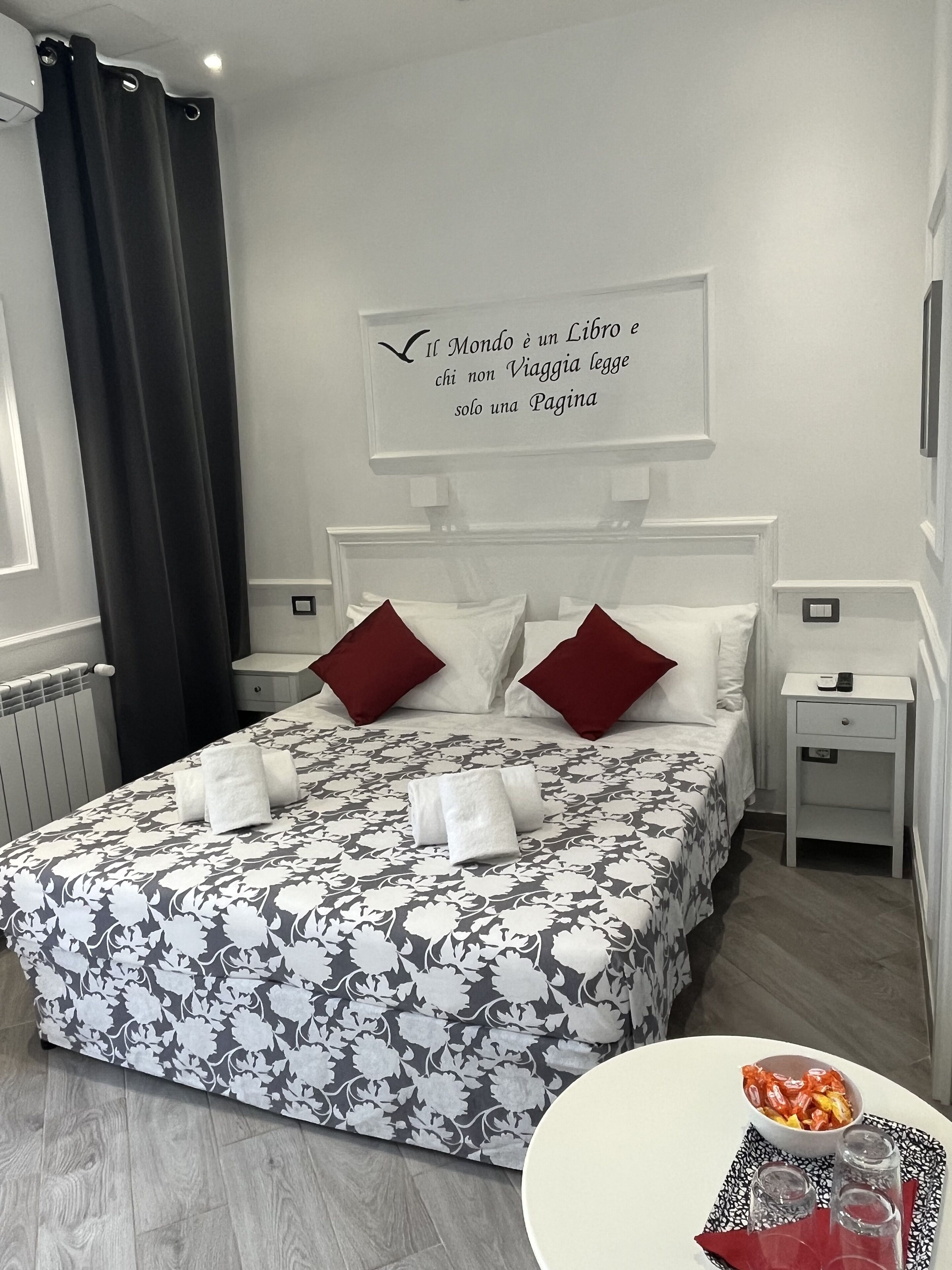 A neatly made bed with a floral bedspread, two red pillows, and a white wall with a quote in Italian.