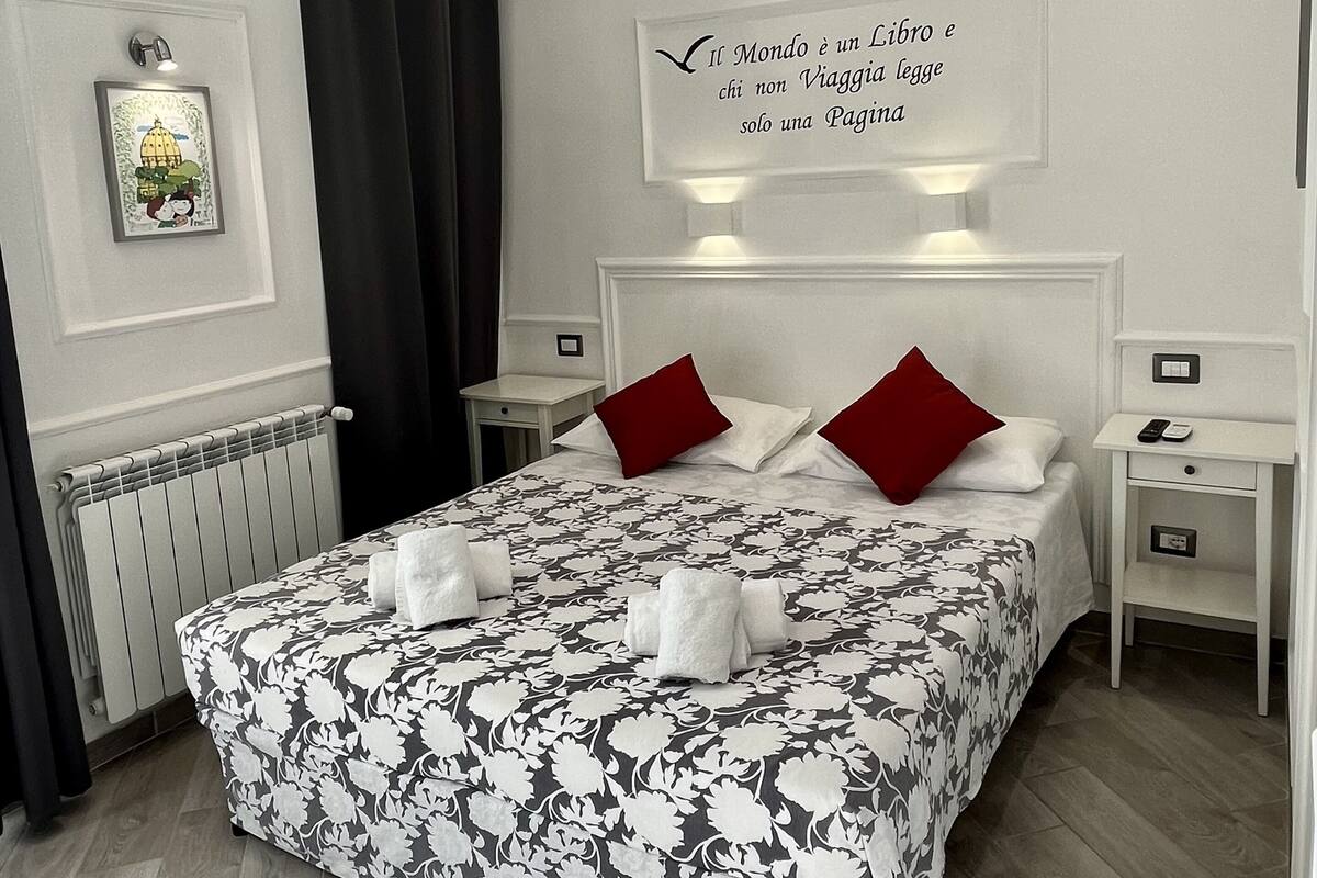 A modern hotel room with a large bed, two red pillows, a white headboard, and a framed quote on the wall.