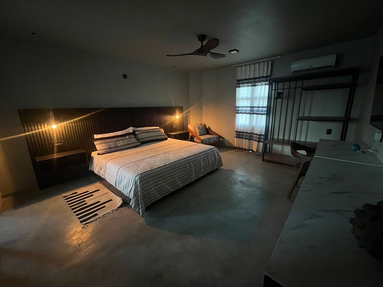 A modern bedroom with a large bed, a desk, a chair, a ceiling fan, and a window with curtains.