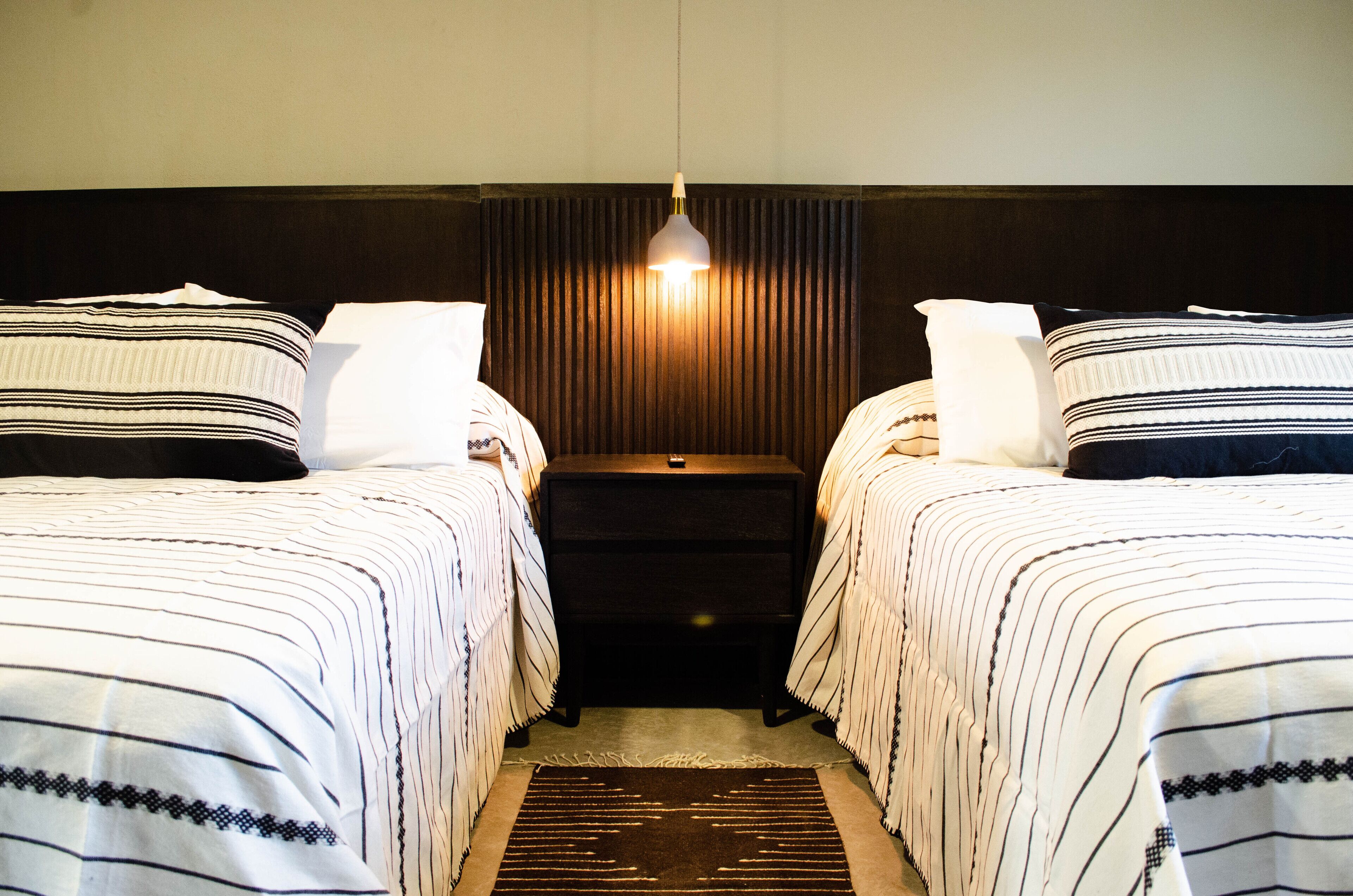 A hotel room with two beds, a dark headboard, a light fixture above the headboard, and a small nightstand between the beds.
