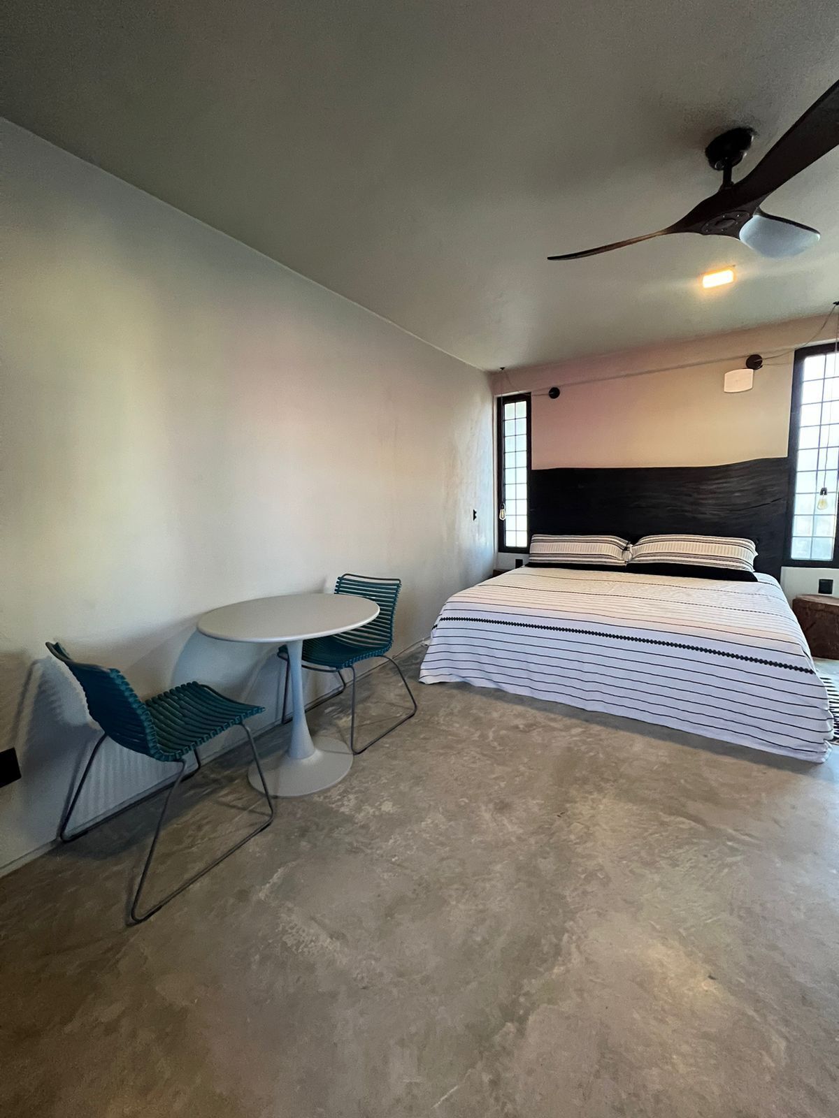 A compact bedroom with a bed, a small table, two chairs, a ceiling fan, and a window.