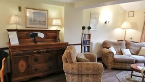 A living room with a wooden piano, two patterned armchairs, a sofa, a bookshelf, and a framed picture on the wall.