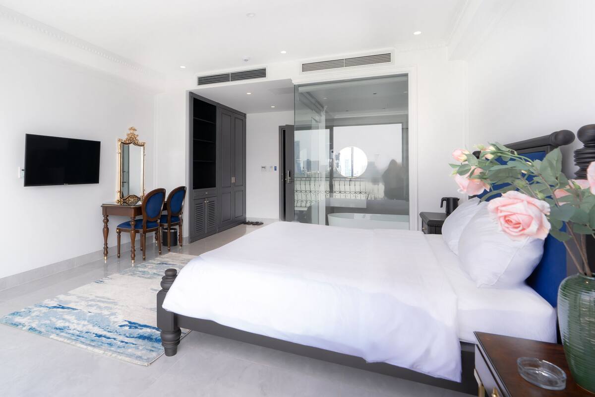 A modern bedroom with a large bed, a TV, a mirror, and a view of the city through a glass door.