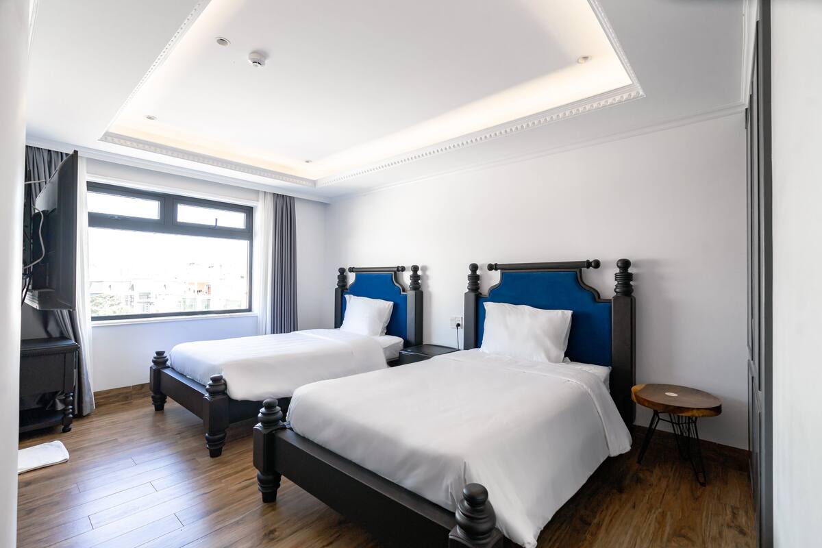 A hotel room with two beds, wooden flooring, a large window, and a small round table.