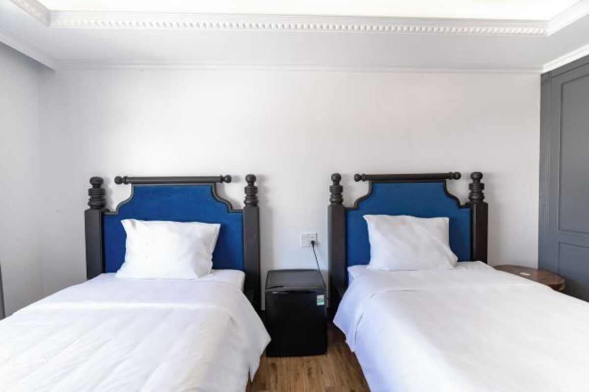 A room with two beds, each with a blue headboard and white bedding. There is a small black speaker on the floor between the beds.