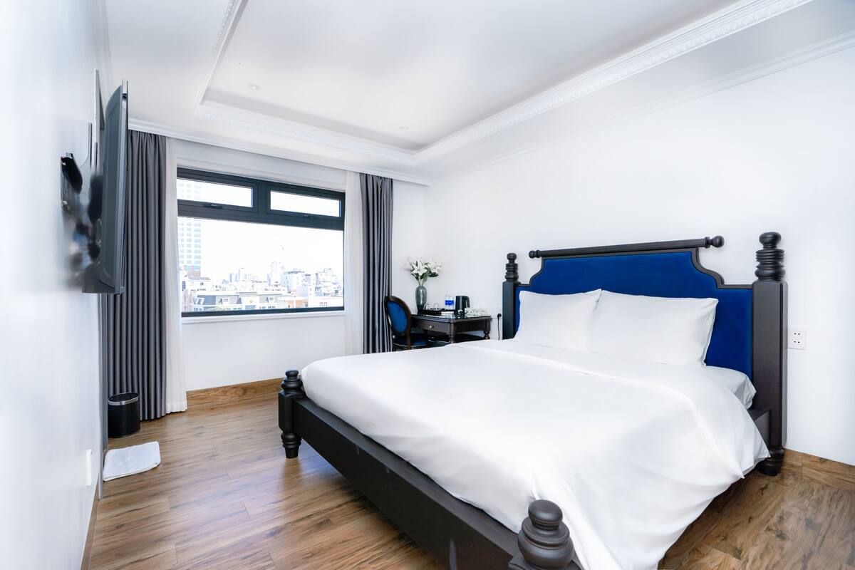 A hotel room with a large bed, a desk, a chair, and a view of the city through a window.