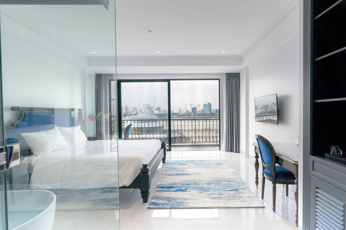 A modern hotel room with a large bed, a glass partition, a city view balcony, and a desk with a chair.