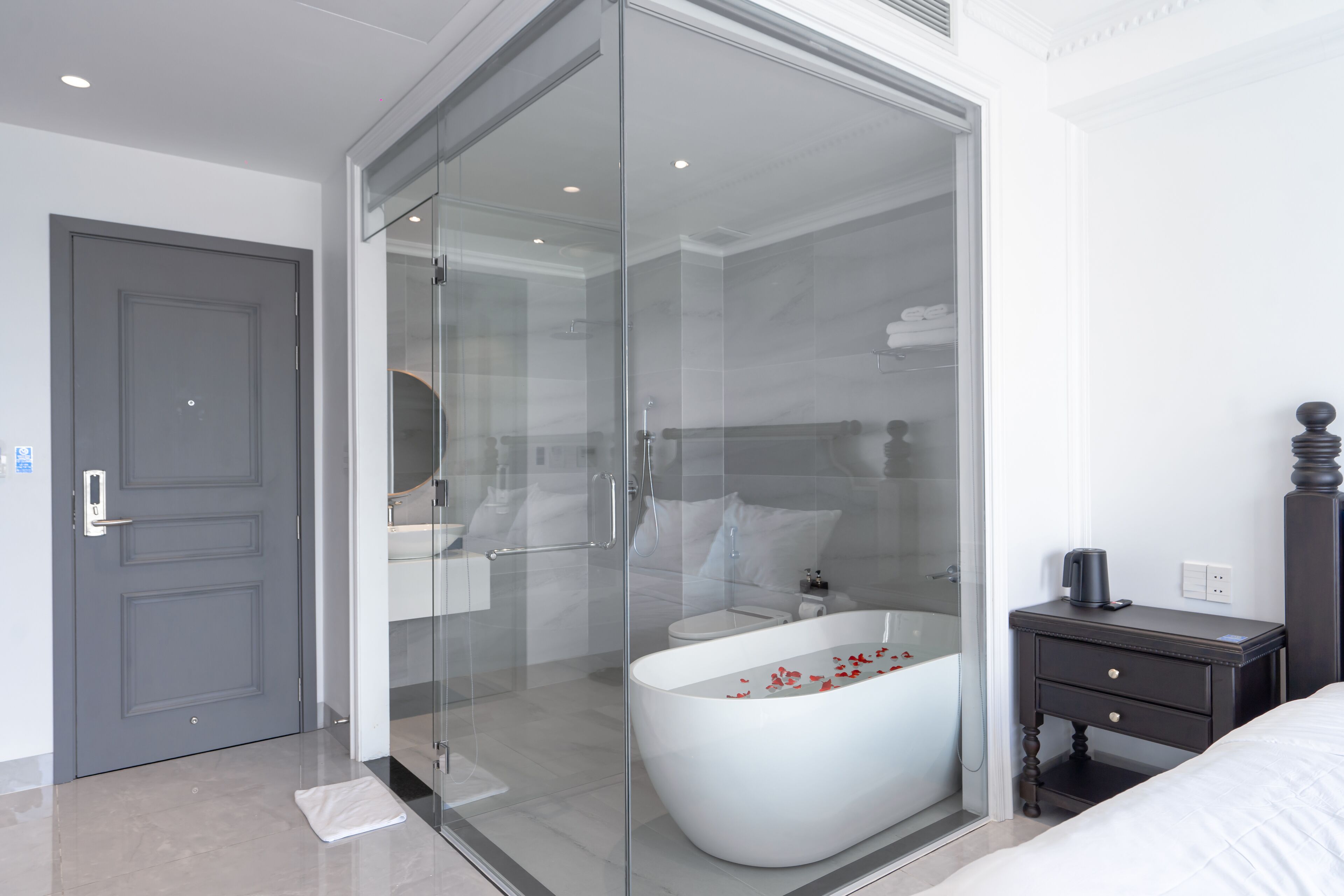 A modern bathroom with a glass-enclosed shower, a freestanding bathtub, and a dark wooden nightstand.