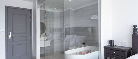 A modern bathroom with a glass-enclosed shower, a freestanding bathtub, and a dark wooden nightstand.