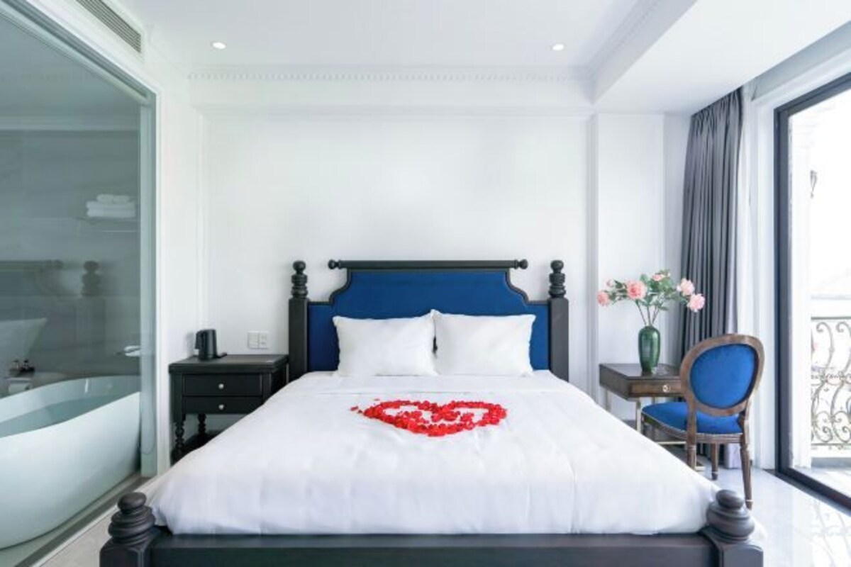 A hotel room with a bed, a nightstand, a chair, a vase with flowers, and a view of the outside.