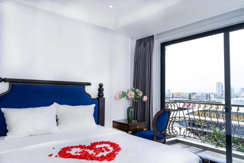 Ein Hotelzimmer mit einem großen Bett, einem Nachttisch, einem Sessel und Blick auf eine Stadt durch große Fenster.
