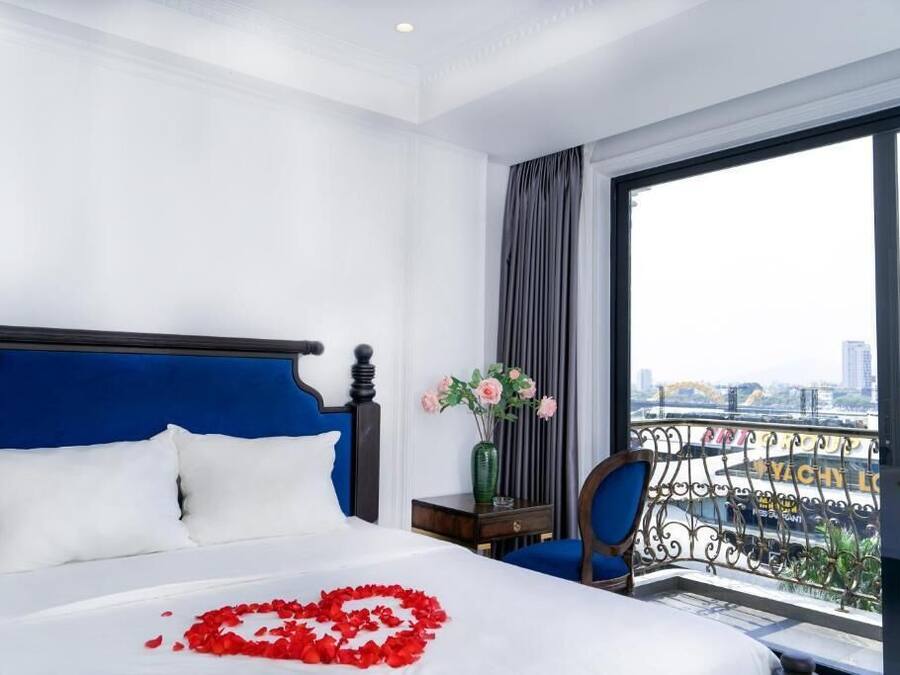 Ein Hotelzimmer mit einem großen Bett, einem Nachttisch, einem Sessel und Blick auf eine Stadt durch große Fenster.