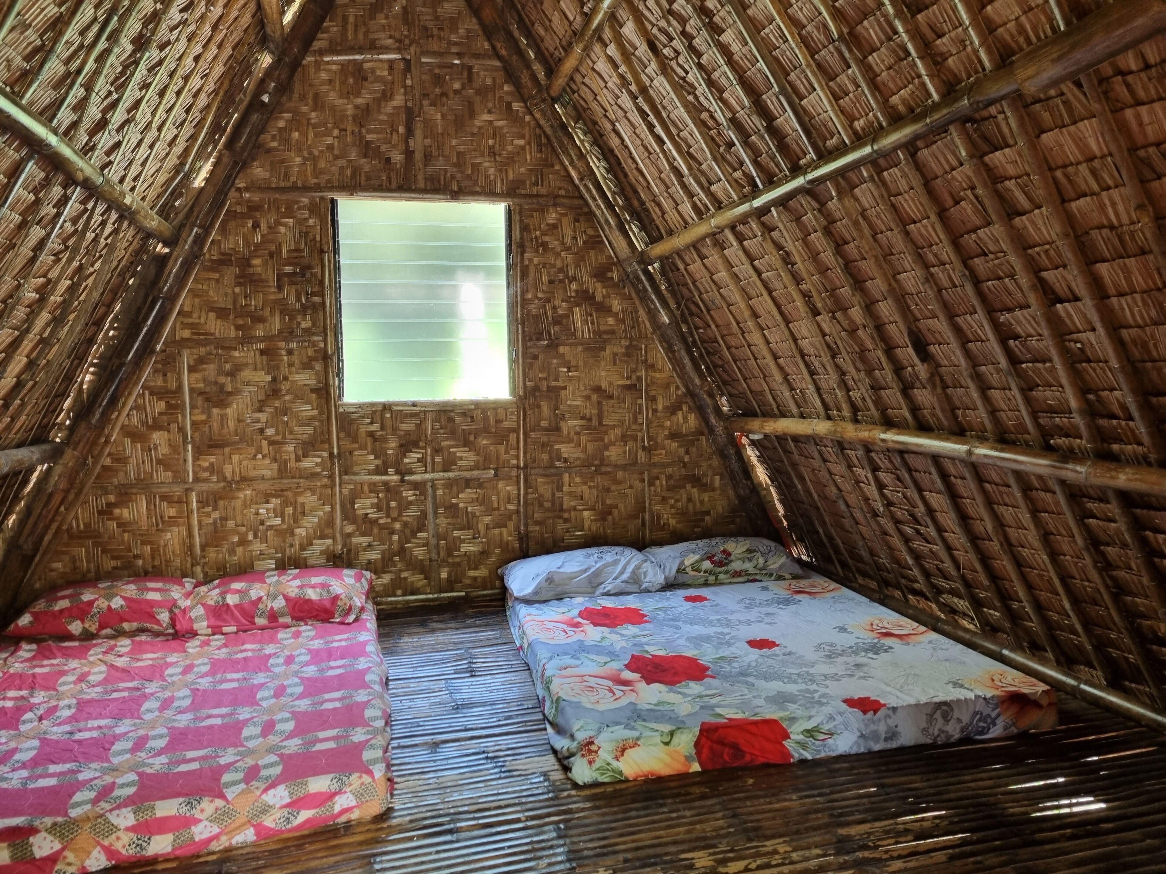 Una habitación con techo de paja, dos camas con ropa de cama floral y una ventana con persianas.