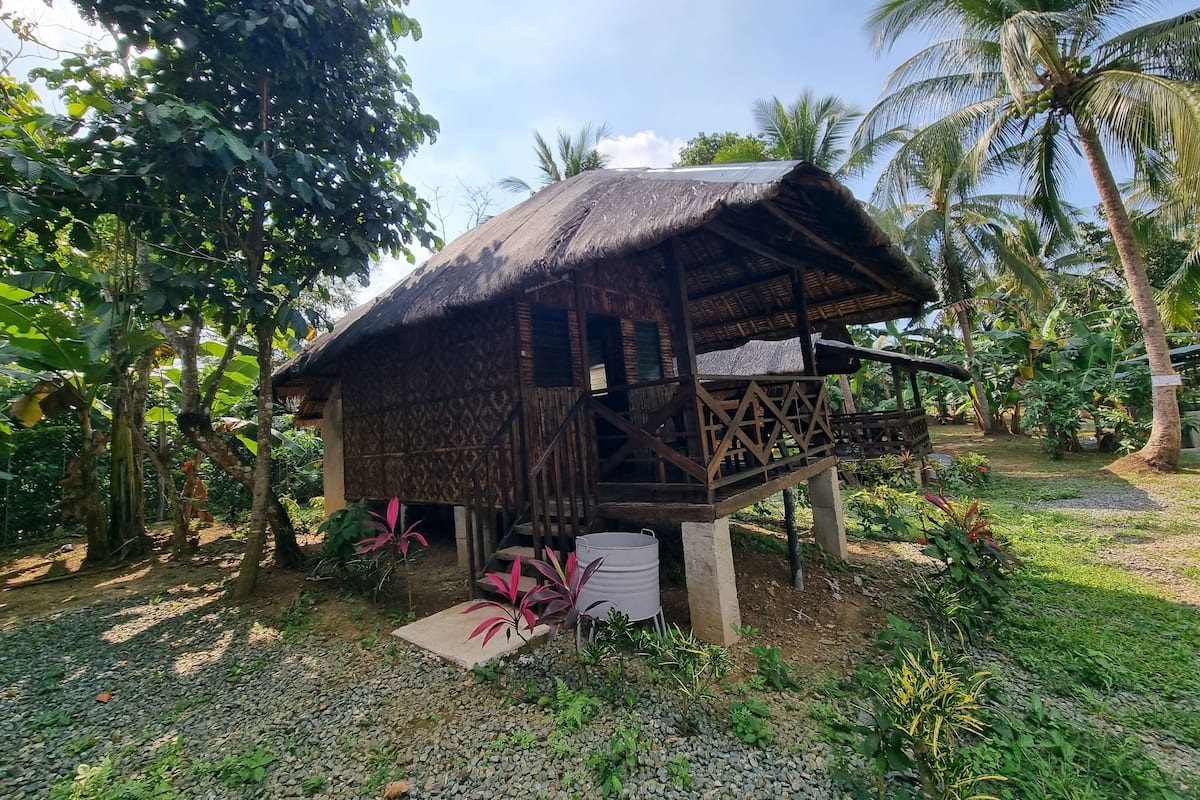 A rustic thatched-roof structure with a balcony, surrounded by tropical vegetation.