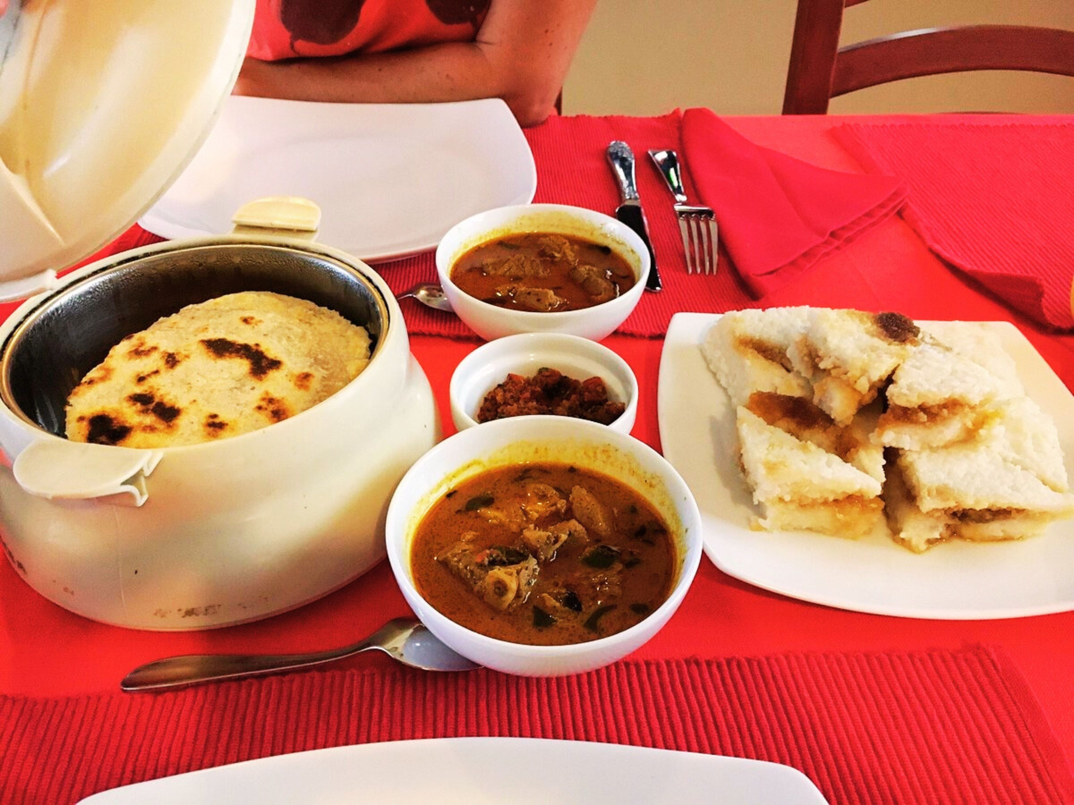 A meal with a pot of rice, two bowls of curry, and a plate of bread on a red tablecloth.