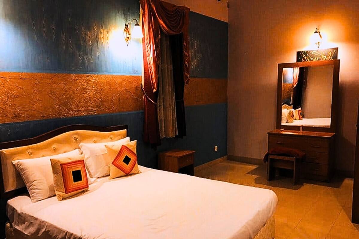 A hotel room with a large bed, a small desk, a mirror, and a window with curtains.