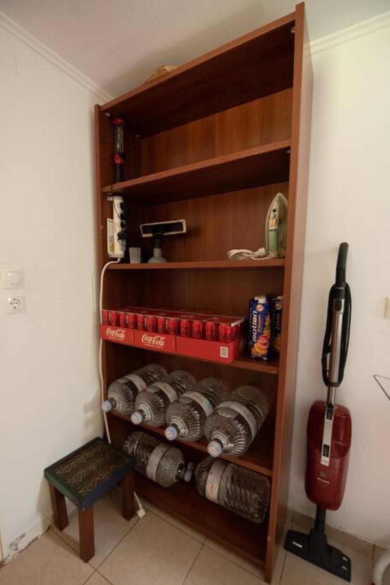 A wooden shelf unit with various items including bottled water, a vacuum cleaner, and a small bench.