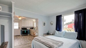 Une chambre avec un lit, une table de chevet, une fenêtre avec des rideaux, une vue sur l’extérieur et, au fond, une petite cuisine.