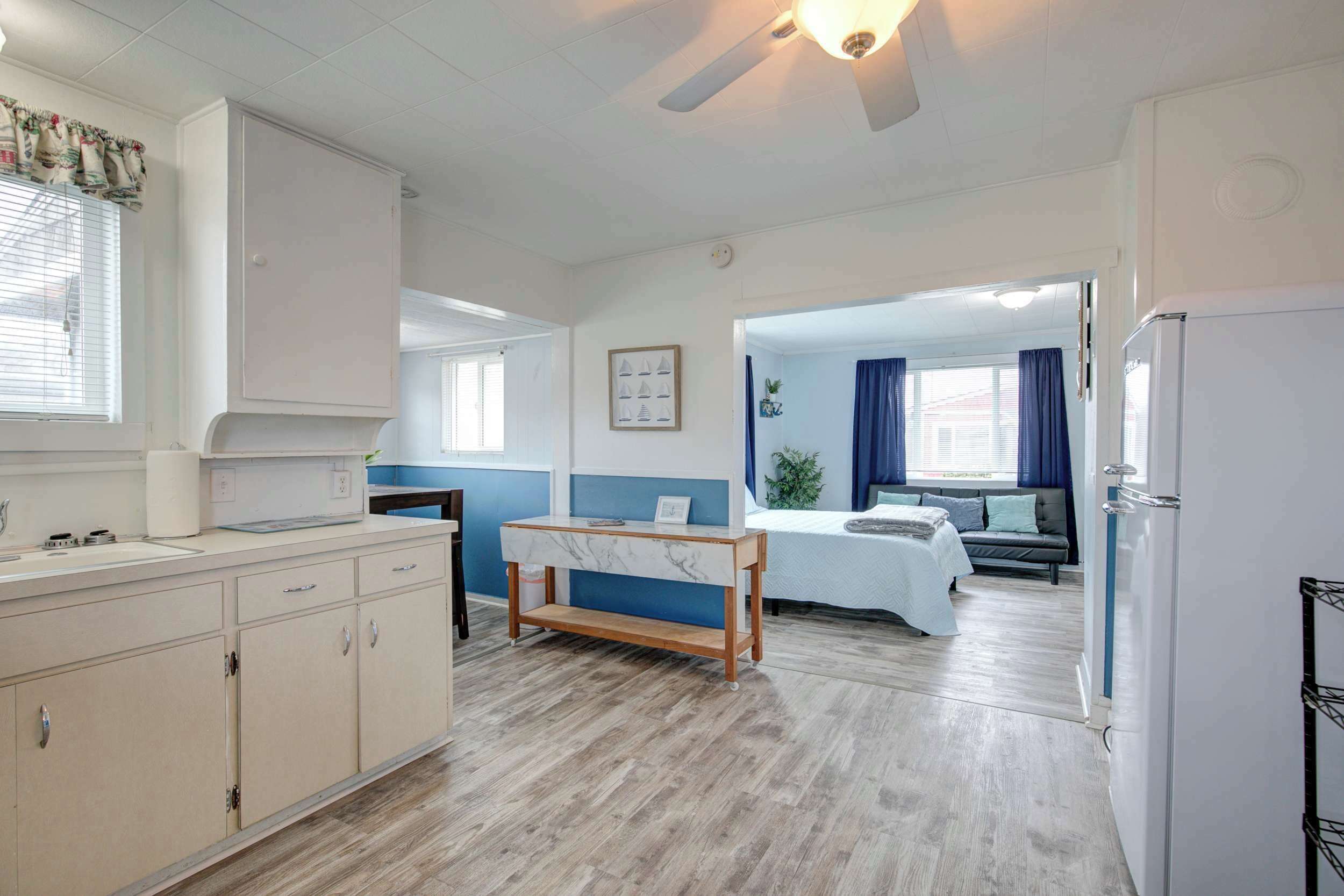 A room with a bed, a desk, a window with curtains, a ceiling fan, and a kitchenette.