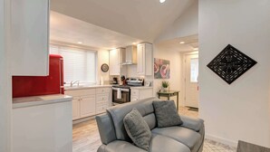 A modern kitchen with white cabinets, a red refrigerator, and a grey sofa with pillows.