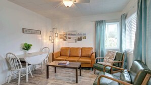 Ein Wohnzimmer mit einer Couch, Sesseln, einem Couchtisch und einem Esstisch. Es gibt einen Deckenventilator, ein Fenster mit Vorhängen sowie Wandbilder.