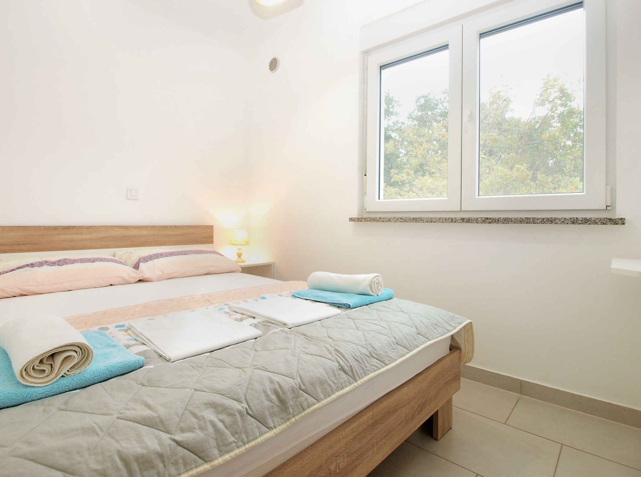 Una cama bien hecha con cubrecama acolchada, una lámpara de noche y una ventana con vistas a árboles.