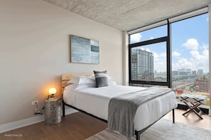 A modern bedroom with a large window, a bed with white bedding, a side table, and a city view.