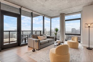 Ein modernes Wohnzimmer mit grauem Sofa, beigen Sesseln, einem Couchtisch und Blick auf die Stadt durch große Fenster.
