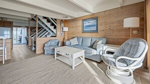 A cozy living room with a wooden wall, a sofa, a coffee table, a wicker chair, and a staircase. - Modern, Spacious Oceanfront Townhome Sleeps 10! (Ocean City)