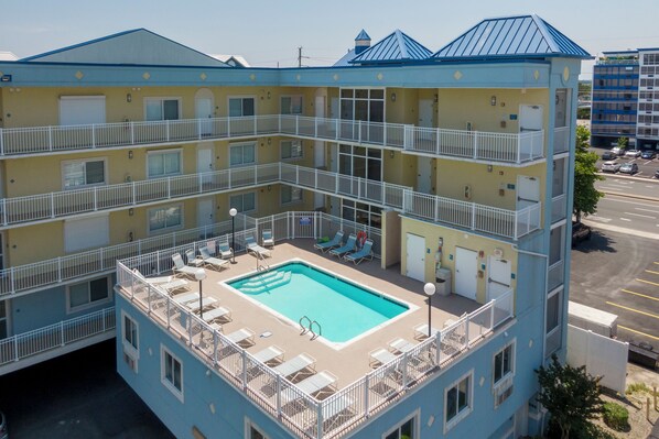 A multi-story hotel with a rooftop pool area, balconies, and a parking lot in the background. - New Listing! Beautiful 4 bedroom oceanblock condo with a pool! (Ocean City)