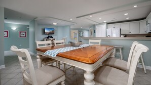 A dining area with a wooden table and white chairs, a kitchen with white cabinets, and a television mounted on the wall.