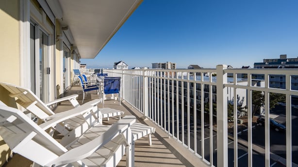 A balcony with white chairs and a blue chair, a white railing, and a clear view of the cityscape.
