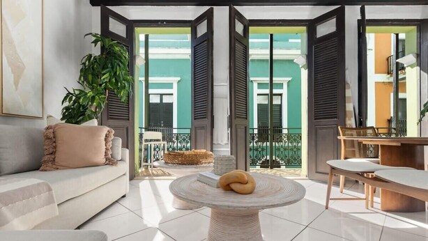 Un salon moderne avec un canapé, une table ronde agrémentée d’un coussin, une plante en pot et une vue sur un bâtiment aux couleurs vives, visible à travers des portes ouvertes.