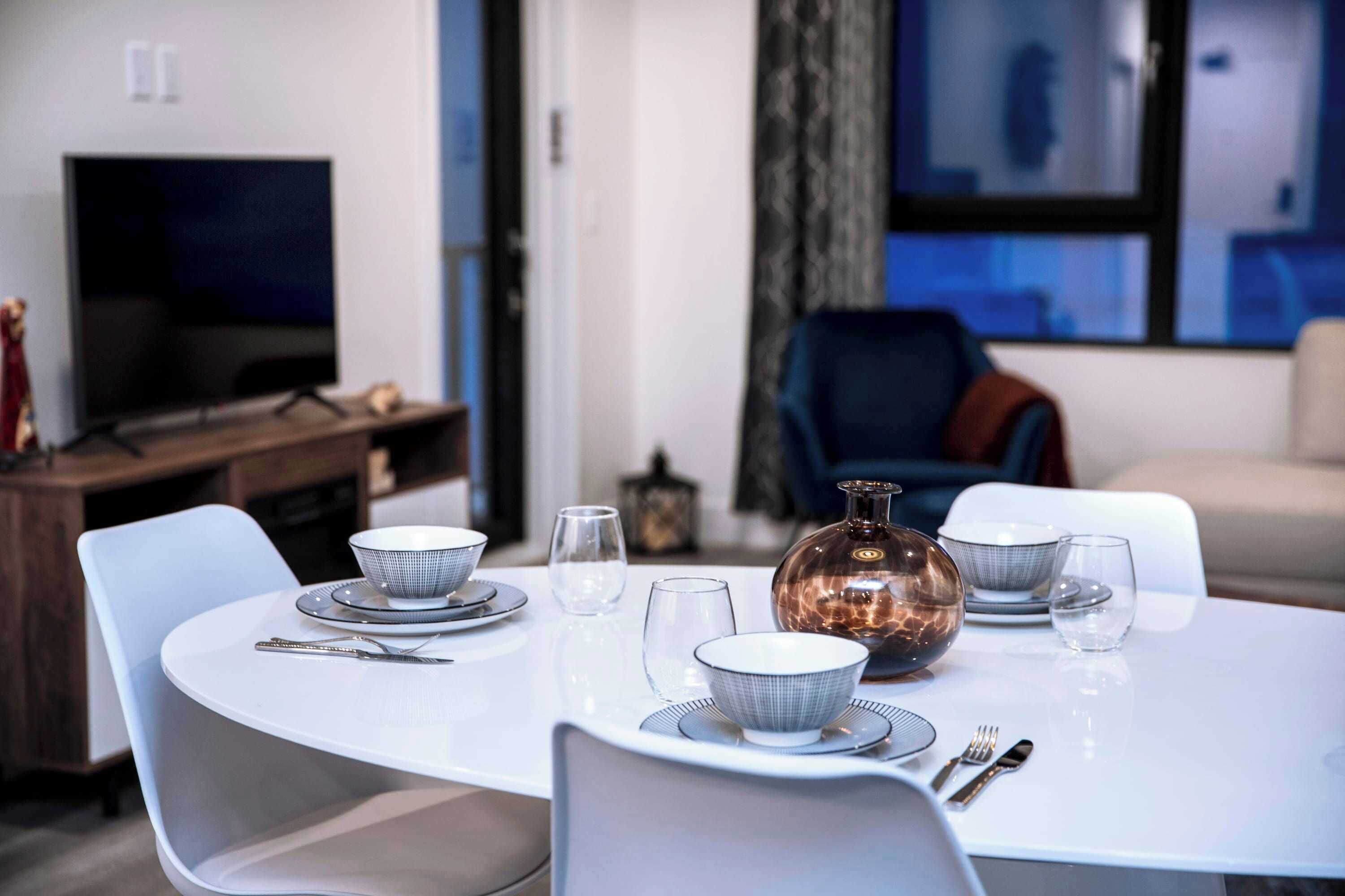 Un comedor con una mesa redonda dispuesta para cuatro personas, un televisor al fondo y vistas de la ciudad a través del ventanal.