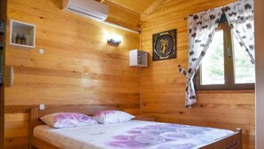 Quarto de cabana de madeira com uma cama, ar condicionado fixado na parede, uma pequena estante com objetos decorativos e uma janela com cortinas.