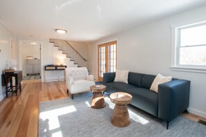 A modern living room with a dark blue sofa, a white armchair, and two wooden side tables. A staircase leads to an adjacent kitchen area. - 5 Min. Drive to Fisher Beach and Pamet Harbor! (Truro)