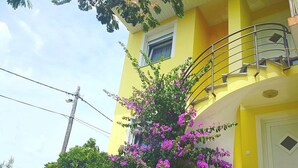 Un bâtiment jaune doté d’un balcon, entouré de verdure et de fleurs violettes.