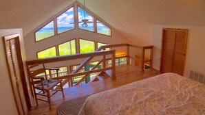 Une chambre avec un grand lit, une chaise en bois, une vue sur l’extérieur par une grande fenêtre et un escalier en bois.