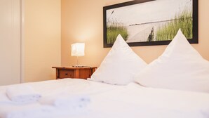 A neatly made bed with white linens and pillows, a bedside table with a lamp, and a framed picture of a beach scene on the wall. - Küstenhostel (Klausdorf)