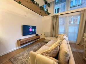 A modern living room with a large flat-screen TV, a sofa, a wooden TV stand, and a window with curtains.