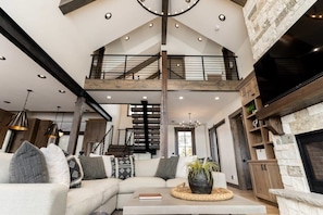 A spacious living area with a high ceiling, a staircase, a fireplace, and a large sofa with cushions. - Lodge at Cutthroat Landing- 20 mins to Yellowstone+Hot Tub+FirePit+Pickleball+LakeAccess+Henry'sLake (Island Park)