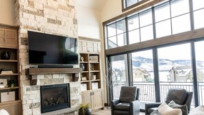 A spacious living room with a stone fireplace, a flat-screen TV, comfortable seating, and large windows offering a view of the outdoors. - Lodge at Cutthroat Landing- 20 mins to Yellowstone+Hot Tub+FirePit+Pickleball+LakeAccess+Henry'sLake (Island Park)