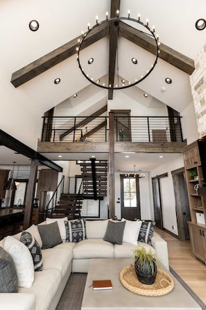 A spacious living area with a high ceiling, exposed wooden beams, a large sectional sofa, and a staircase leading to an upper level. - Lodge at Cutthroat Landing- 20 mins to Yellowstone+Hot Tub+FirePit+Pickleball+LakeAccess+Henry'sLake (Island Park)