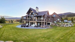 A large, multi-story house with a spacious patio area, surrounded by a well-maintained lawn and a mountainous backdrop. - Lodge at Cutthroat Landing- 20 mins to Yellowstone+Hot Tub+FirePit+Pickleball+LakeAccess+Henry'sLake (Island Park)