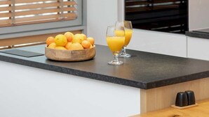 Una cocina moderna con un bol de frutas de madera, dos vasos de zumo de naranja y una ventana con persianas.