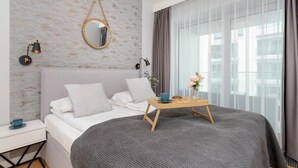 Un dormitorio moderno con una cama grande, una mesita de noche con lámpara, un espejo en la pared y una ventana con cortinas.