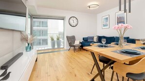 Un comedor moderno con una mesa de madera dispuesta para cuatro, un sillón gris y un sofá azul. Se ve un reloj de pared grande, y hay cuadros enmarcados en la pared.