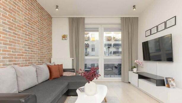 A modern living room with a brick wall, grey sofa, white coffee table, and a TV mounted on the wall. - Elegant Apartment with Balcony | Close to the Airport (Warszawa)