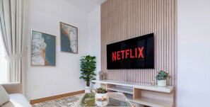 Un salon moderne avec une table basse en verre, un canapé blanc, un téléviseur fixé au mur et deux œuvres d’art encadrées.