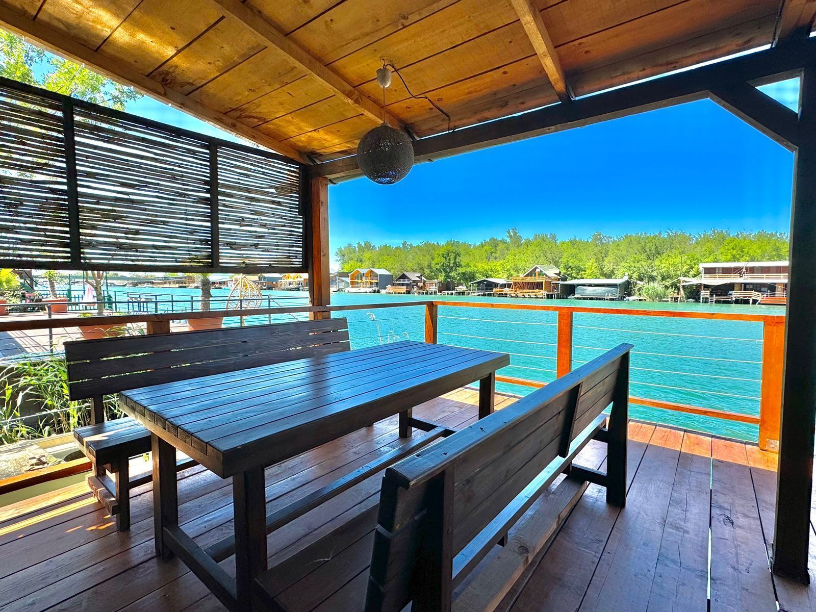Eine Holzterrasse mit Tisch und Bänken, mit Blick auf ein Gewässer und Häuser im Hintergrund.